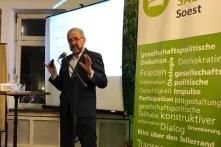Das Bild zeigt Reinhard Loske (bärtig, grauhaarig, Anzug, gestikulierend hinter einem Standmikrofon vor einer weißen Wand) während seines Vortrags beim Grünen Salon Soest
