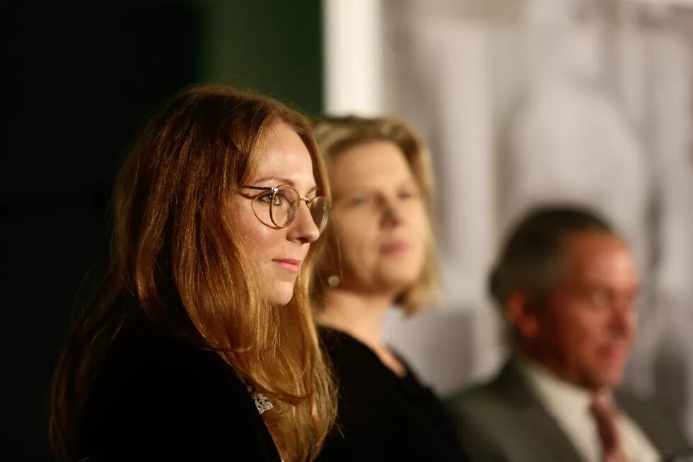 Das Bild zeigt eine junge, rootharige Frau mit Brille, unscharf im Hintergrund zwei weitere Personen