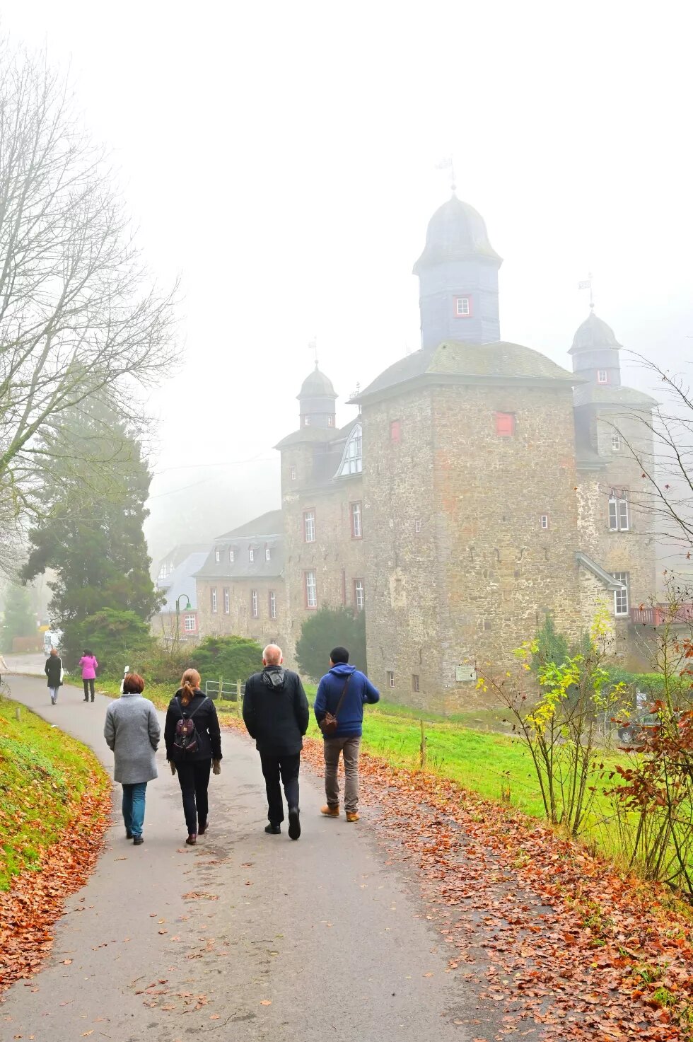 Das Bild zeigt ein Schloss im Nebel, vor dem vier Personen in herbstlicher Kleidung mit dem Rücken zur Kamera auf einem Weg stehen