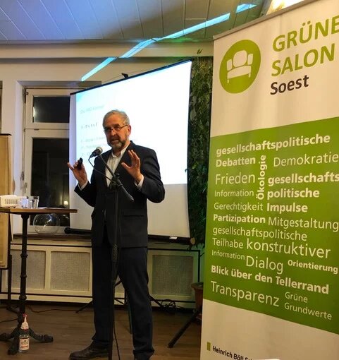 Das Bild zeigt Reinhard Loske (bärtig, grauhaarig, Anzug, gestikulierend hinter einem Standmikrofon vor einer weißen Wand) während seines Vortrags beim Grünen Salon Soest