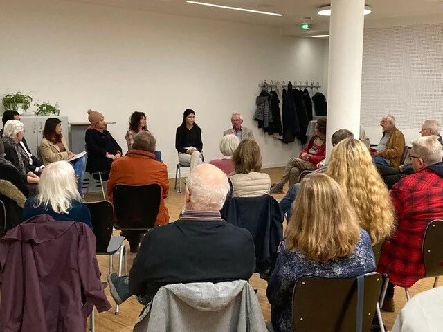 Eine Gruppe von Menschen, die im Stuhlkreis sitzen und einer Person zuhören