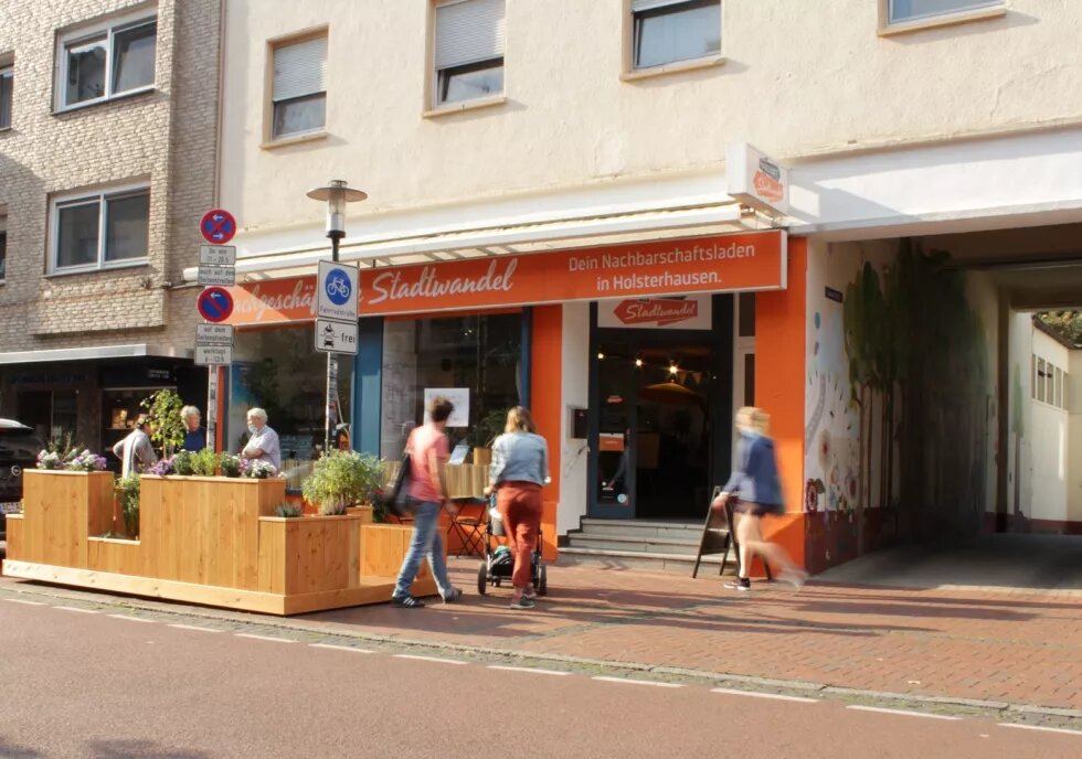 Das Bild zeigt die Außenfront des Fachgeschäfts für Stadtwandel in Essen-Holsterhaußen, ein orangefarbenes Schafenster, Menschen laufen vorbei.