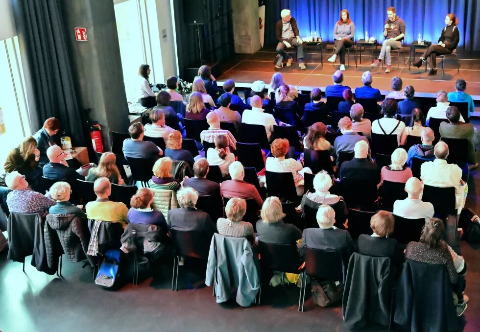 Das Bild zeigt ca. 60 Menschen von hinten, die auf Stühlen vor einer Bühne sitzen, auf der vier Personen sitzen.