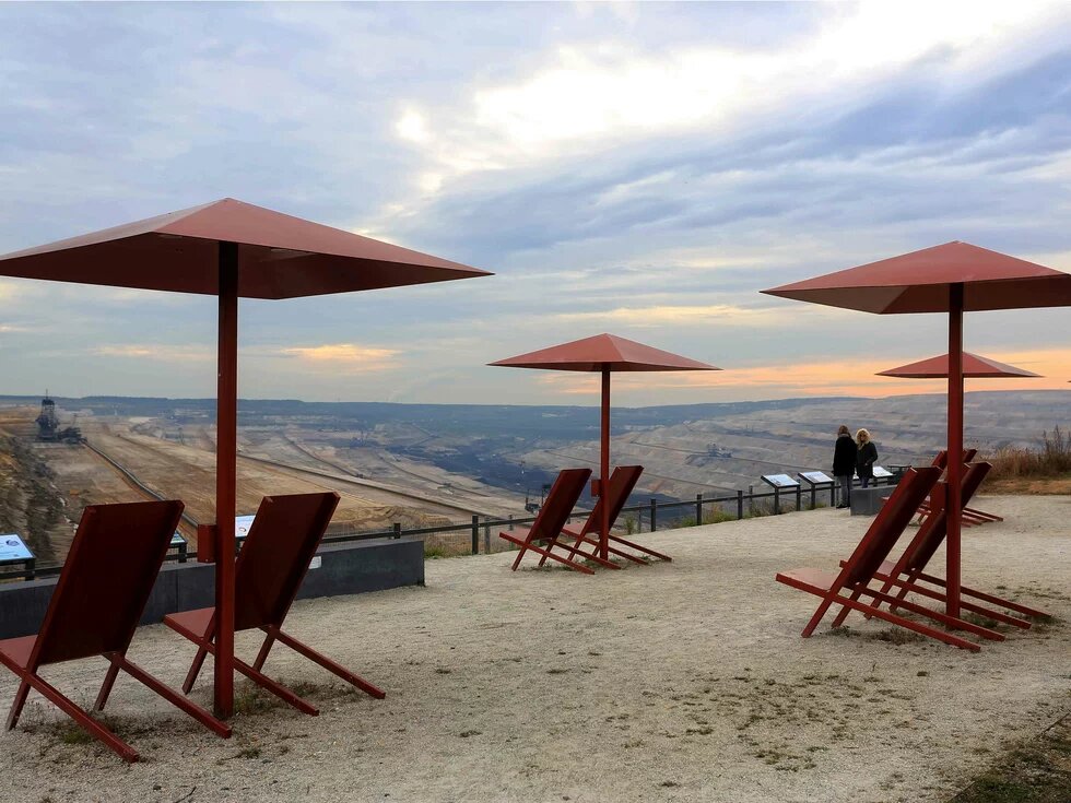 Das Bild zeigt einen Panoramablick auf ein Braunkohlegebiet, im Vordergrund Sonnenstühle und Schirme auf einer Terrasse.