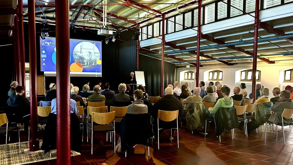 Das Bild zeigt Menschen auf Stühlen bei einer Veranstaltung, die einer Bühne zugewandt sind.