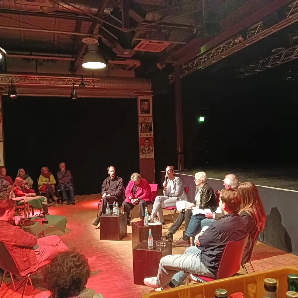 Die Teilnehmenden der Diskussion sitzen im Halbkreis vor den Zuschauenden. 